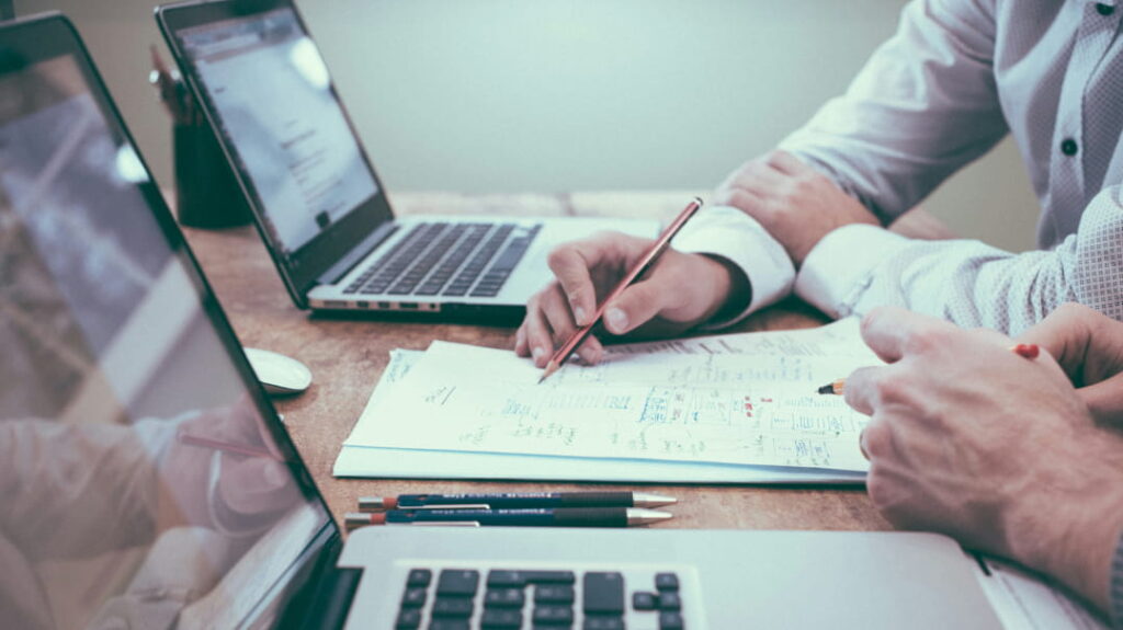 Two people reviewing financial documents at a desk with laptops, discussing planning and advice
